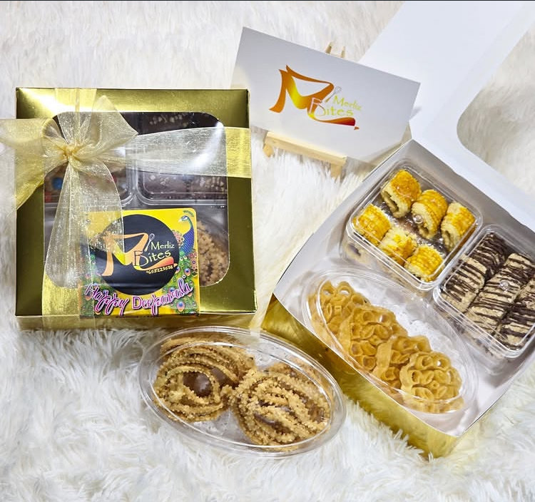 A gold Deepavali gift box tied with organza ribbon, beside an open box of achi muruku, festive bites and chocolate-drizzled bars